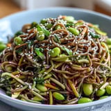 Overhead view of a Soba Noodle Bowl drizzled with sesame dressing, garnished with fresh herbs on a rustic table.